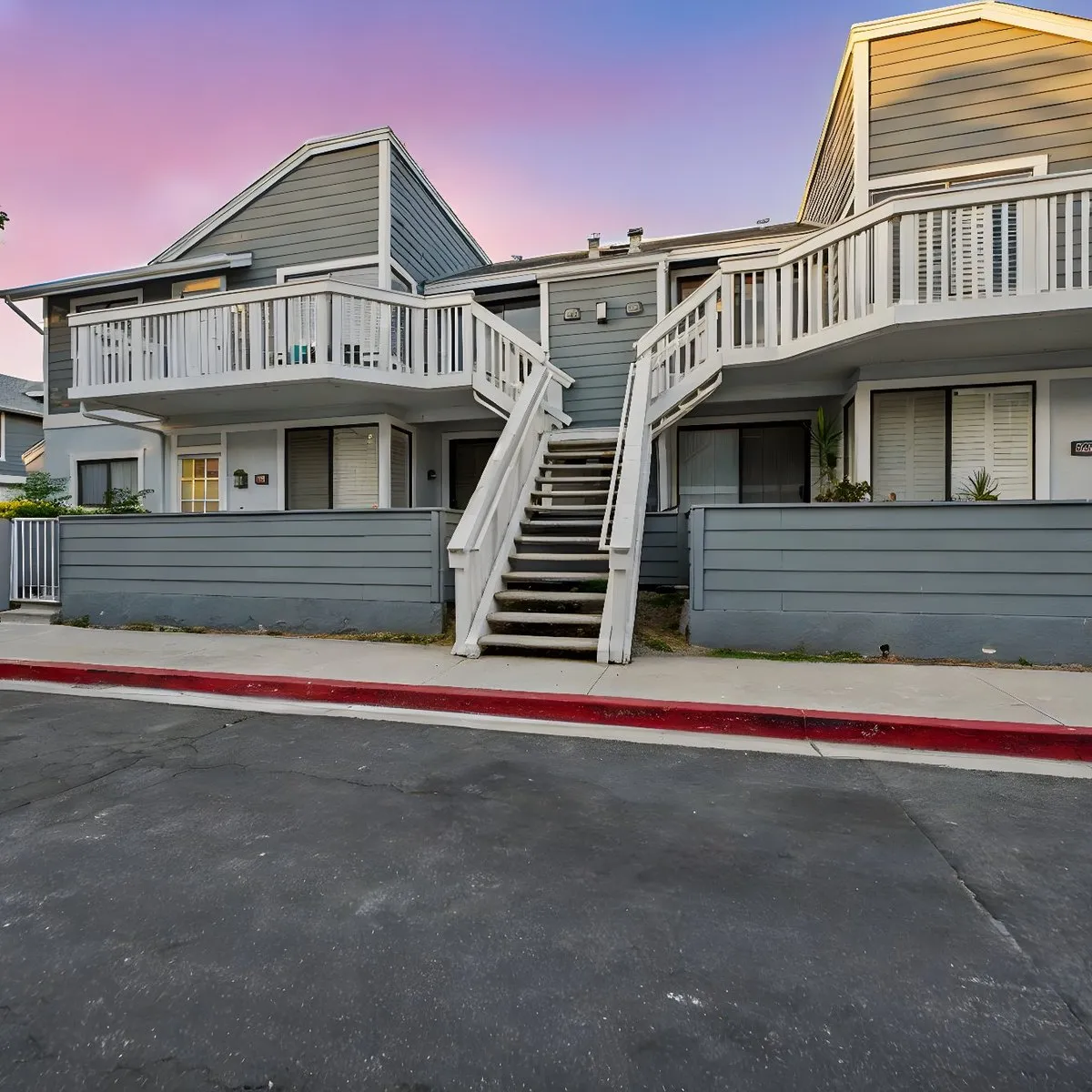 A two-story gray condo building with white balconies and a center staircase under a pink evening sky.